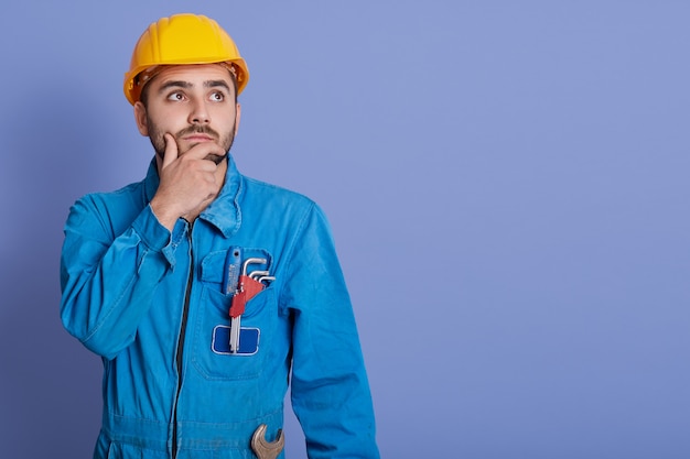 Pensive guy wearing yellow helmet and blue overalls, boy with wrench, looking aside with thoughtful facial expression