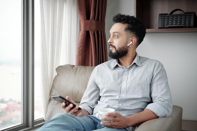 Pensive handsome bearded man sitting in armchair listening to music in earbuds drinking coffee and l...