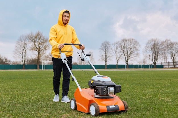 Pensive handsome guy in yellow hoodie and black pants with gasoline mower in his hands mows grass on lawn