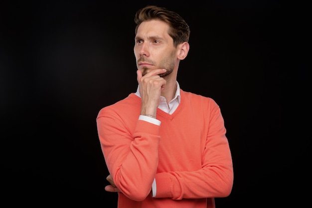 Pensive handsome young man in orange sweater rubbing chin while thinking of strategy, black background