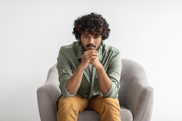 Pensive indian guy sitting in armchair over white