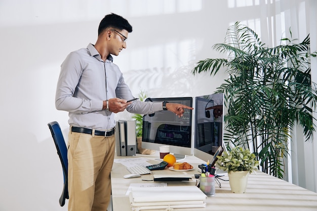 Pensive Indian programmer with smartphone in hand pointing at computer screen with programming code on it