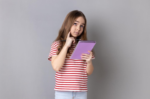 Pensive little girl wearing striped Tshirt writing in organizer making to do list