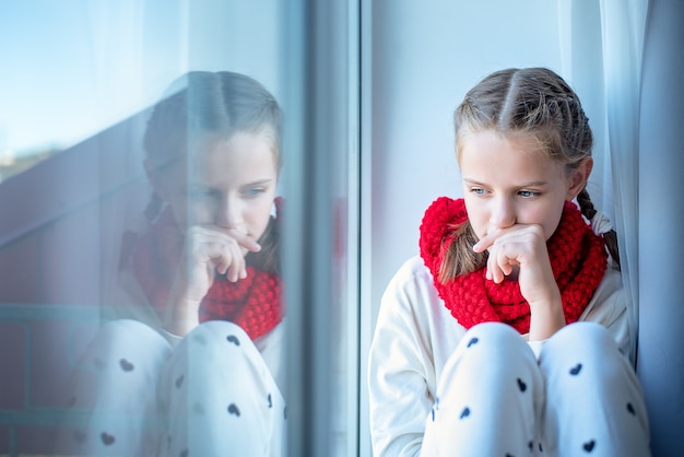 A pensive little girl with a scarf around her neck sits on the windowsill and is reflected in the window. Loneliness concept.