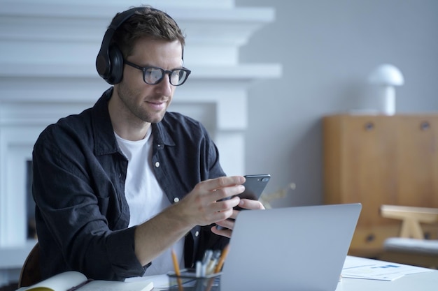 Pensive male entrepreneur in headphones sitting at desk at home office and using mobile phone