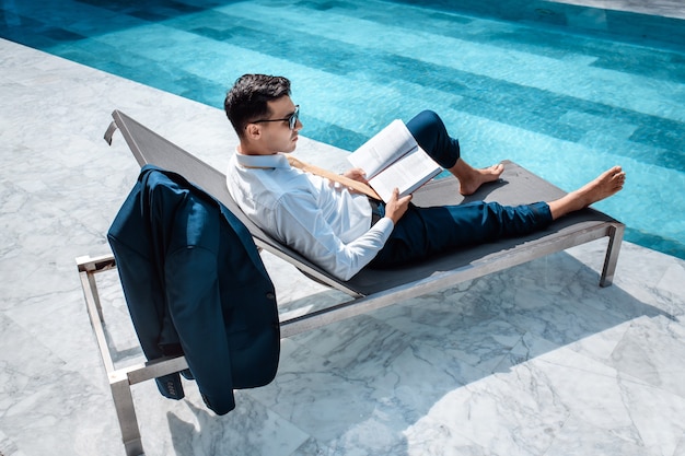 A pensive man in a business suit is resting near the pool with a book in his hands on a lounger