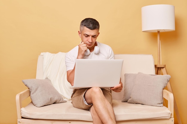 Pensive man wearing white Tshirt and shorts sitting on sofa against beige background working on notebook thinking new job ideas holding chin being concentrated