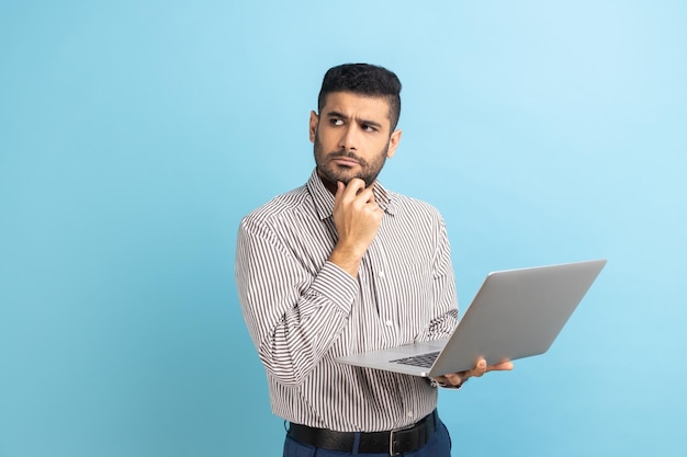 Pensive man with beard holding laptop and holding chin looking away thinking over business project