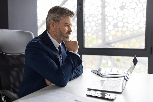 Pensive Middle Aged Businessman Working With Laptop At Desk In Office