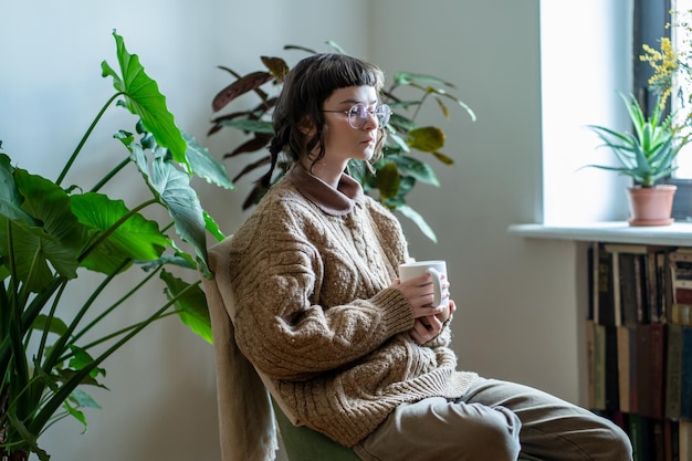 Pensive millennial girl sitting with phone and cup of tea in urban jungle home looking out window