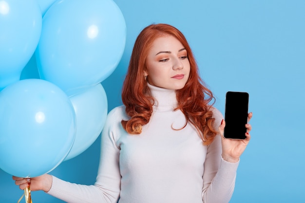 Pensive red haired European woman thinks celebrate birthday holiday party, holds modern mobile phone with blank screen and bunch of helium balloons in hands, wears white turtleneck, female looks away.