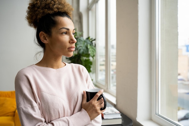 Pensive sad africanamerican woman standing at the window with a cup of coffee in her hands