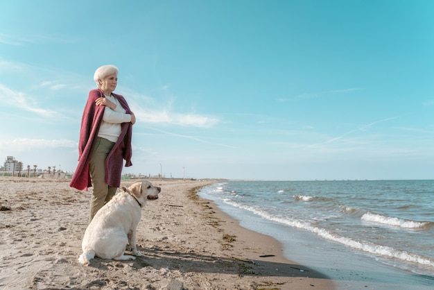 Pensive senior lady and a cute dog standing on the sandy beach by the sea