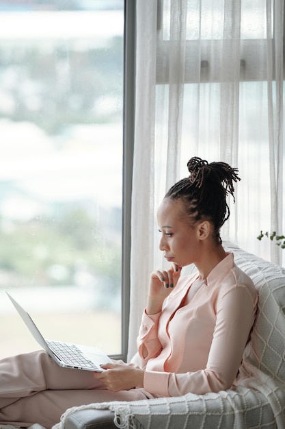 Pensive serious female entrepreneur in pajamas reading document on laptop screen when working at home