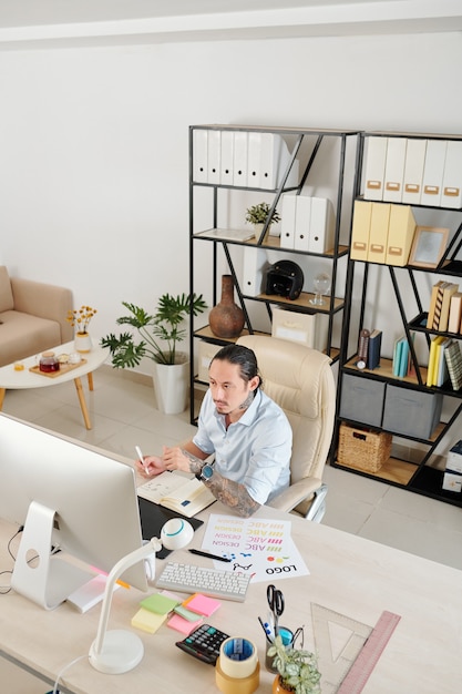 Pensive serious graphic designer working at home office, checking e-mail on computer screen and taking notes in planner