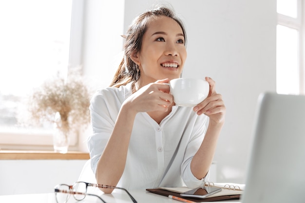 Pensive smiling asian business woman drinking coffee and looking away while sitting by the table in office