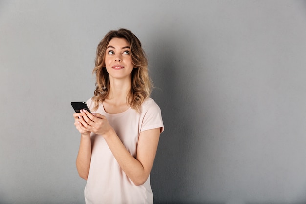 Pensive smiling woman in t-shirt holding smartphone and looking up over grey bacground