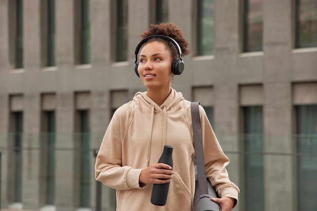 Pensive sporty curly woman dressed in beige hoodie strolls outdoors against modern building listens music via headphones holds bottle of water carries rolled karemat leads healthy lifestyle