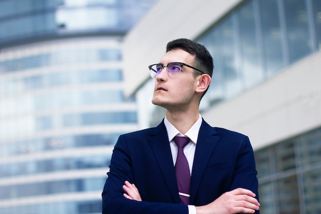 Pensive thoughtful businessman in suit, tie and glasses standing outdoor in front of office modern building