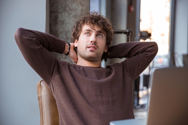 Photo pensive thoughtful dreaming curly attractive handsome man in brown sweetshirt holding hands behind head while sitting at his working place