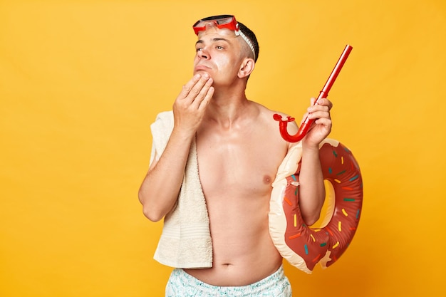 Pensive thoughtful tourist man at summer resort standing with rubber ring isolated over yellow background posing with cream on face holding chin thinking