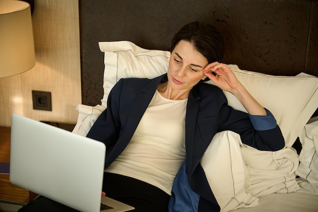 Pensive tired beautiful Caucasian business woman in elegant casual suit working on laptop lying on the bed, resting after hard day at work