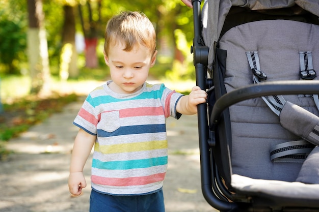 Pensive toddler boy stands near the stroller in the park Cute kid of one and a half years Selective focus