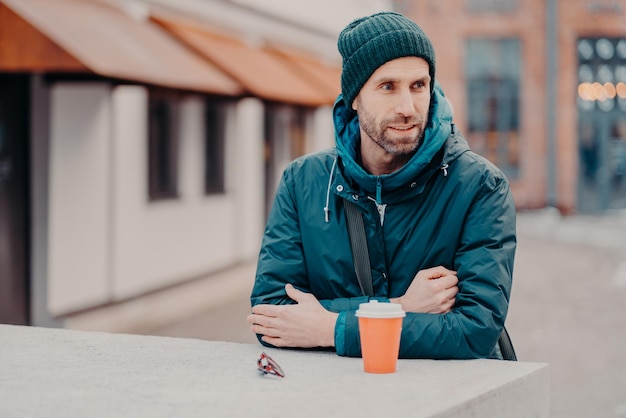 Pensive unshaven young Caucasian man with bristle wears hat and jacket with hoody drinks takeaway coffee stands outdoor has stroll People lifestyle spare time and street style concept