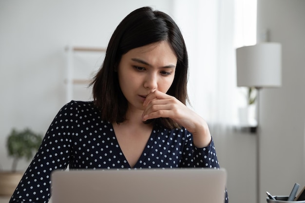 Pensive Vietnamese young woman look at laptop screen think of work problem solution or consider idea Thoughtful Asian millennial girl study on computer browse internet make decision pondering