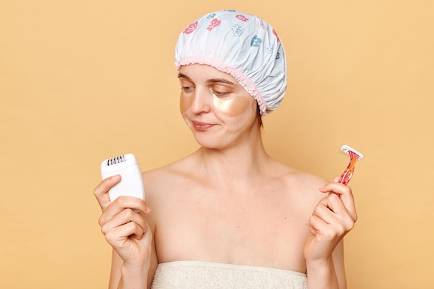 Pensive woman wearing shower cap standing isolated over beige background taking care of her armpit skin choosing razor or epilator