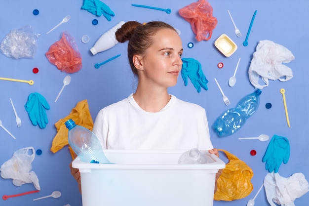 Pensive woman wearing white shirt isolated on blue wall with litter around holding can with rubbish looking away trash contaminates earth recycling reduces waste pollution