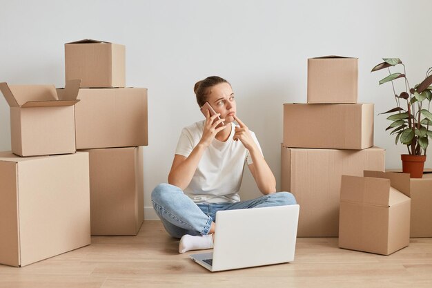 Pensive woman with hair bun wearing white T-shirt sitting on the floor near cardboard boxes, talking phone with delivery service, looking away, thinking about new interior during relocating.