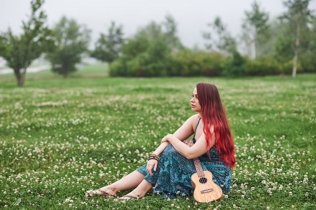 Pensive woman with red hair sitting on the grass in the summer park, holding a ukulele.