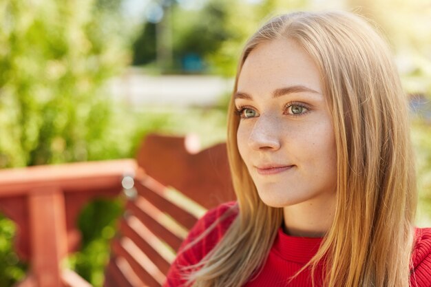 Pensive woman with sad appealing eyes, freckled pure skin, round face and blonde hair looking aside while sitting at bench in park analyzing what she done and dreaming about something good in her life