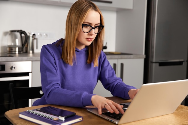 Pensive young adult woman in glasses work study at kitchen using laptop remote education