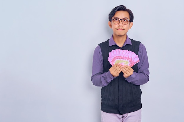 Pensive young Asian man in purple shirt clothes holding cash money in rupiah banknotes and looking away at copy space isolated on white background People lifestyle concept