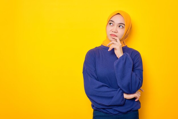 Pensive young Asian Muslim woman dressed in casual sweater looking up at copy space and touching chin thinking about interesting offer isolated over yellow background