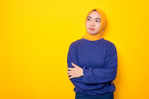 Pensive young Asian Muslim woman dressed in casual sweater looking up at copy space and touching chin thinking about interesting offer isolated over yellow background