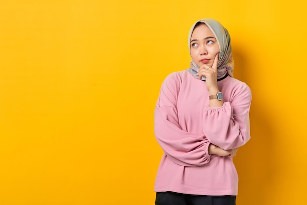 Pensive young Asian woman in pink shirt looks seriously thinking about a question on yellow background