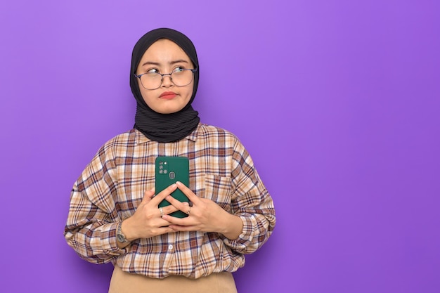 Pensive young Asian woman in plaid shirt holding a mobile phone looking away at copy space thinking about question isolated on purple background