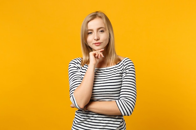 Pensive young blonde woman in striped clothes put hand prop up on chin isolated on bright yellow orange wall background, studio portrait. People sincere emotions lifestyle concept. Mock up copy space.