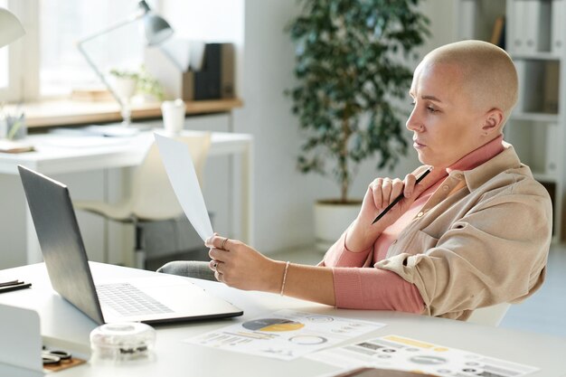 Pensive young Caucasian businesswoman with bald hairstyle sitting at desk with papers and laptop and analyzing document