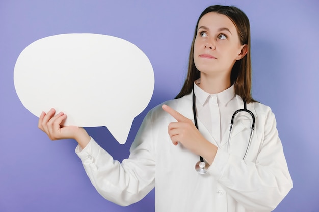 Pensive young doctor woman in medical gown stethoscope hold empty blank Say cloud speech bubble, posing isolated over violet studio background wall. Healthcare personnel health medicine concept