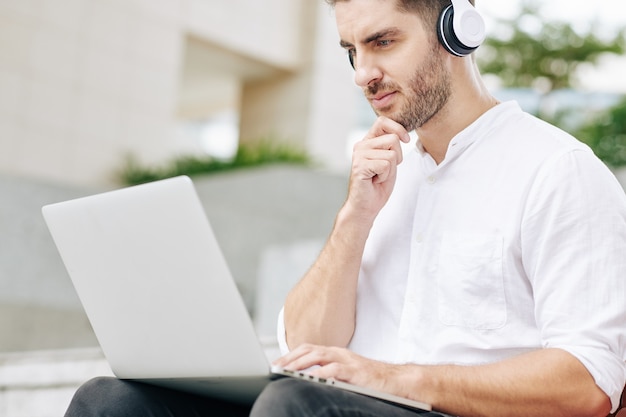 Pensive young freelance software developer rubbing chin and looking at laptop screen