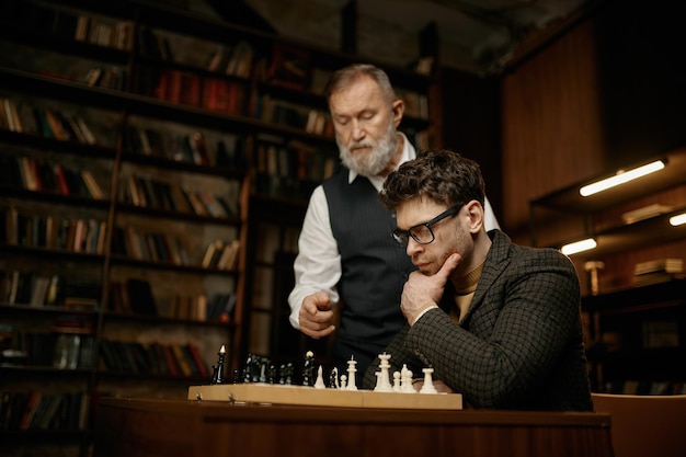 Pensive young man looking at chess board and listening hints older father. Senior parent teaching son playing strategic game at home library. Weekend intelligent activity concept