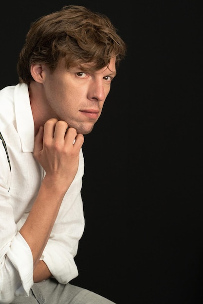 Pensive young man sitting on chair and looking at camera isolated on black background close up portrait