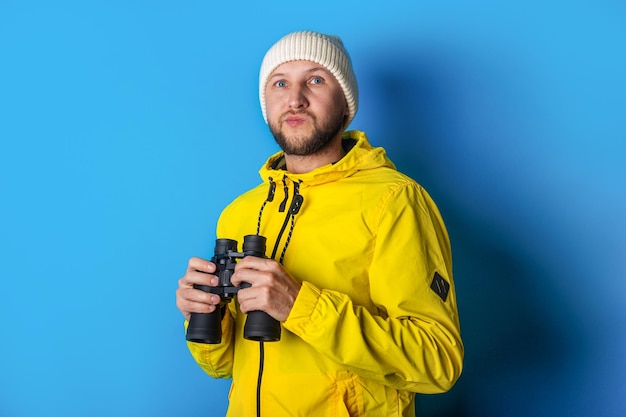 Pensive young man in a yellow jacket with binoculars on a blue background