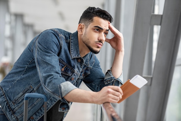 Pensive young middle eastern man with passport and tickets standing in airport