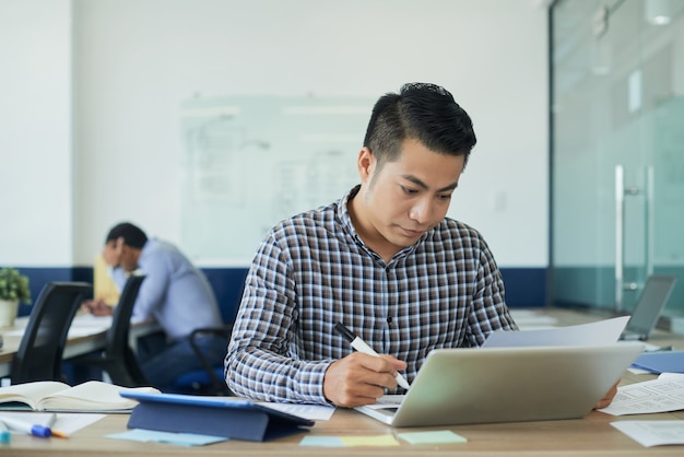 Pensive young UI designer working at office desk with opened laptop and checking document with layout of website interface