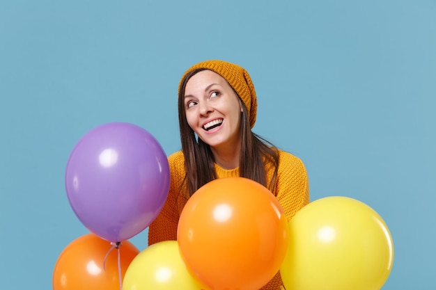 Pensive young woman girl in sweater and hat posing isolated on blue background. Birthday holiday party, people emotions concept. Mock up copy space. Celebrating hold colorful air balloons looking up.